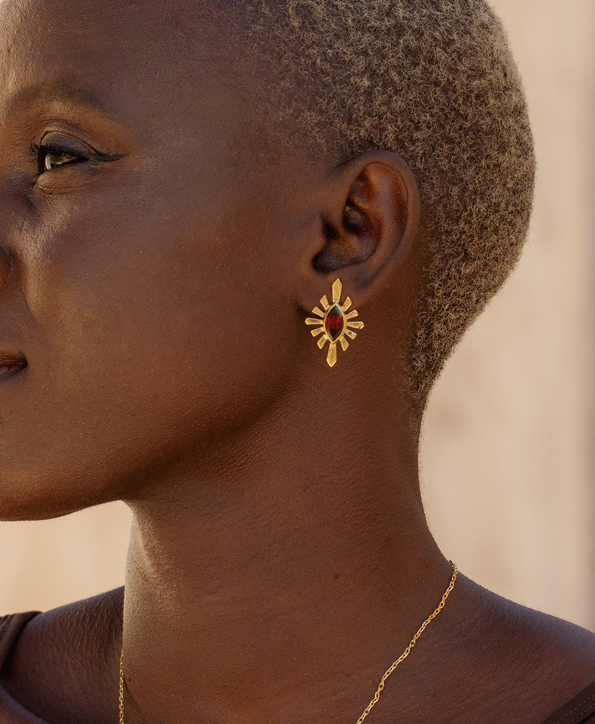 A close-up side profile of a person wearing the Sister Studs, featuring a marquise cut garnet in a gold linear sunburst frame. 