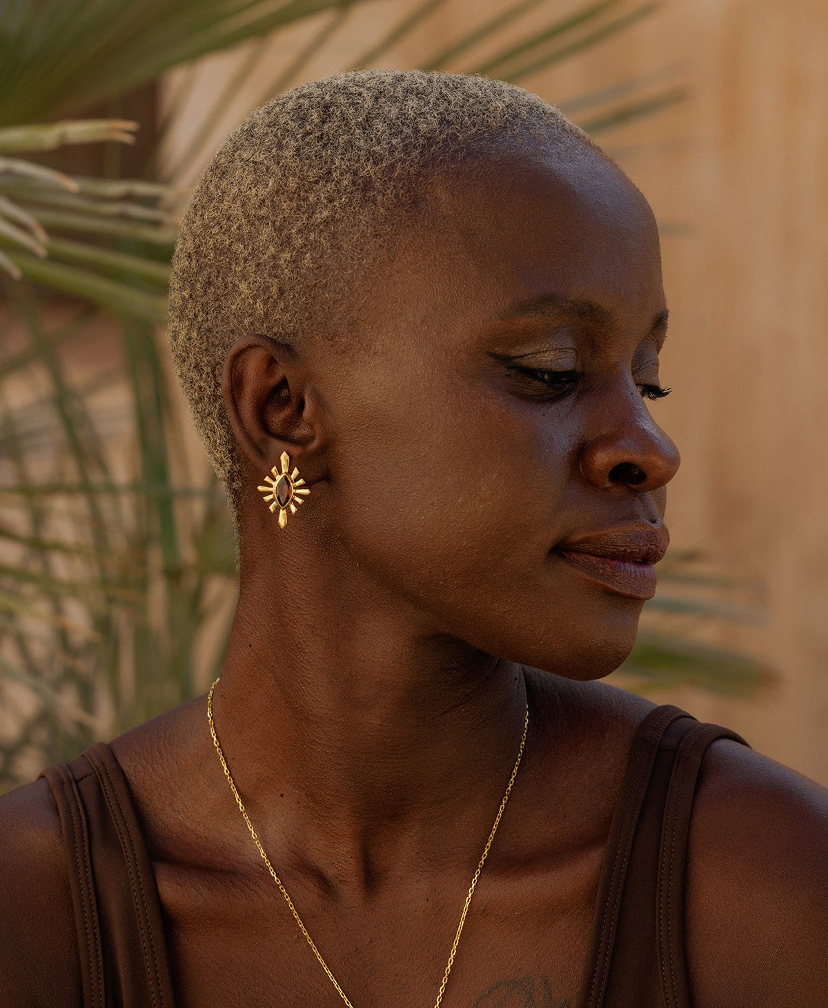 A side profile of a person wearing the Sister Studs, featuring a marquise cut garnet in a gold linear sunburst frame. 