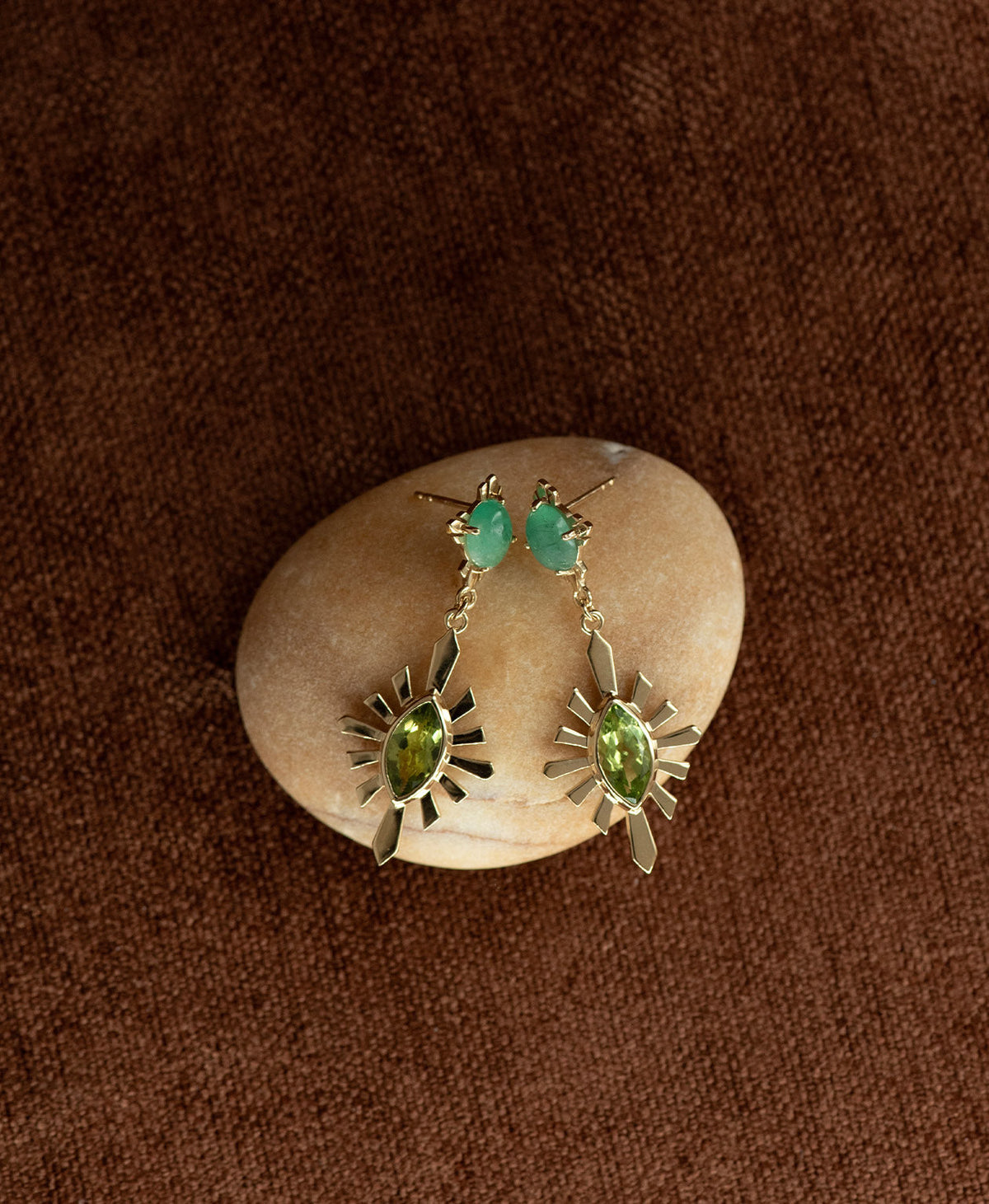 A pair of gold Sisterhood Earrings with a cabochon cut Emerald, set within a linear border, paired with a marquise cut Peridot nestled with a sunburst frame, displayed on a smooth beige stone against a textured brown fabric background.