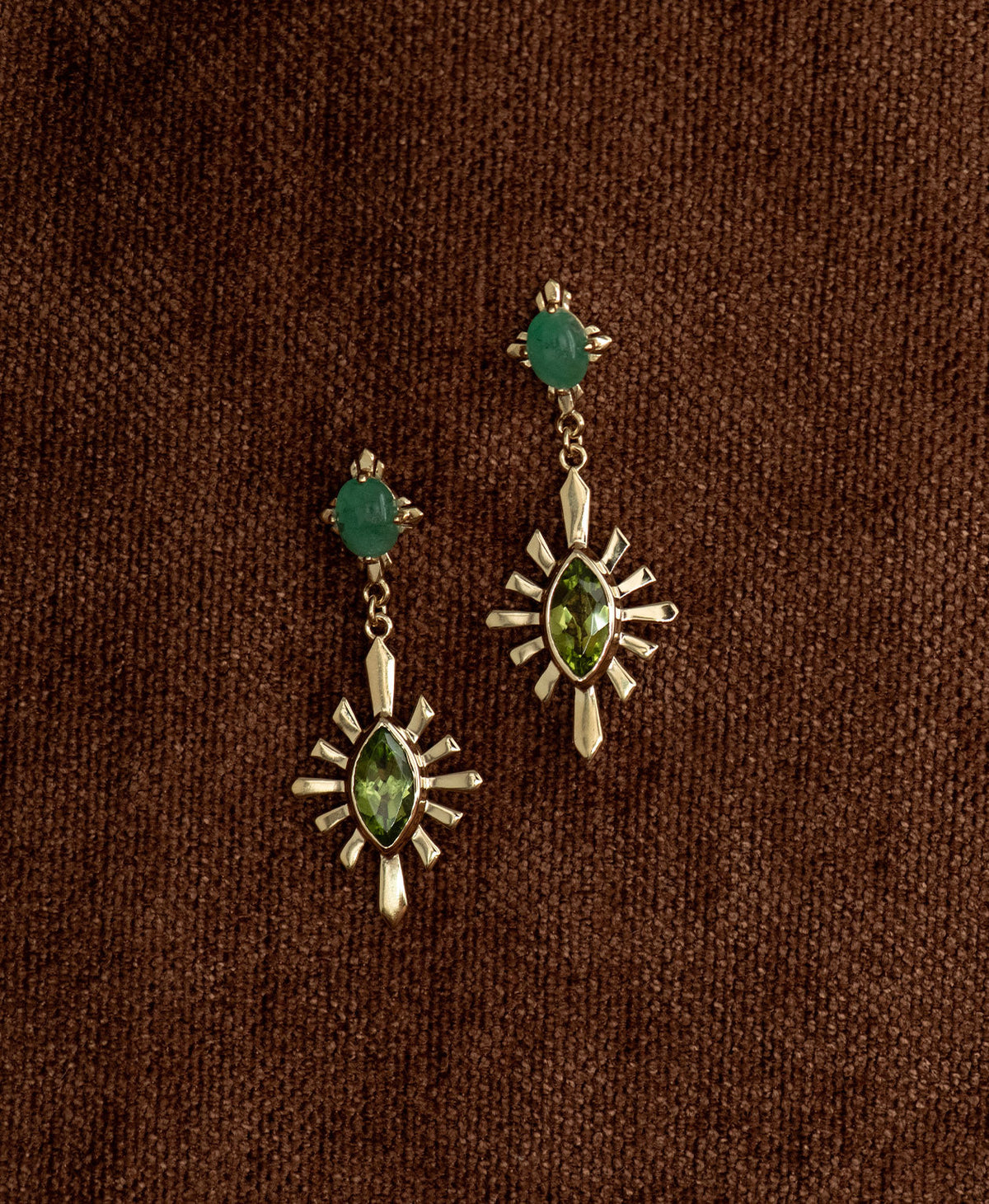 A pair of gold Sisterhood Earrings with a cabochon cut Emerald, set within a linear border, paired with a marquise cut Peridot nestled with a sunburst frame, hanging against a textured brown fabric background.