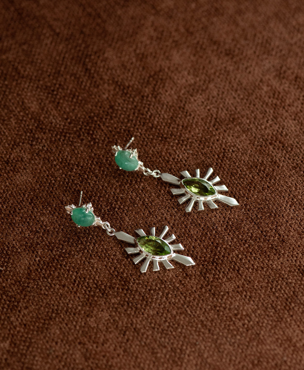 A pair of silver Sisterhood Earrings with a cabochon cut Emerald, set within a linear border, paired with a marquise cut Peridot nestled with a sunburst frame, displayed against a textured brown fabric background.