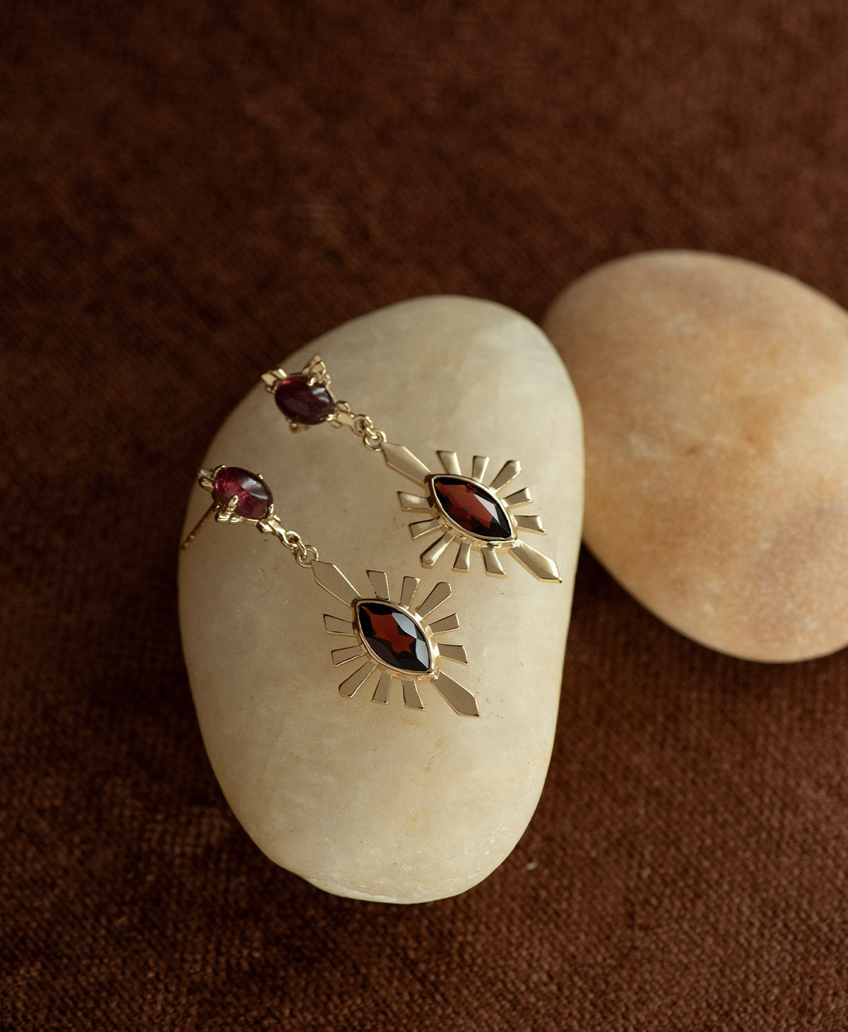 A pair of gold Sisterhood Earrings with a cabochon cut pink Tourmaline, set within a linear border, paired with a marquise cut Garnet nestled with a sunburst frame, displayed on a smooth beige stone against a textured brown fabric background.