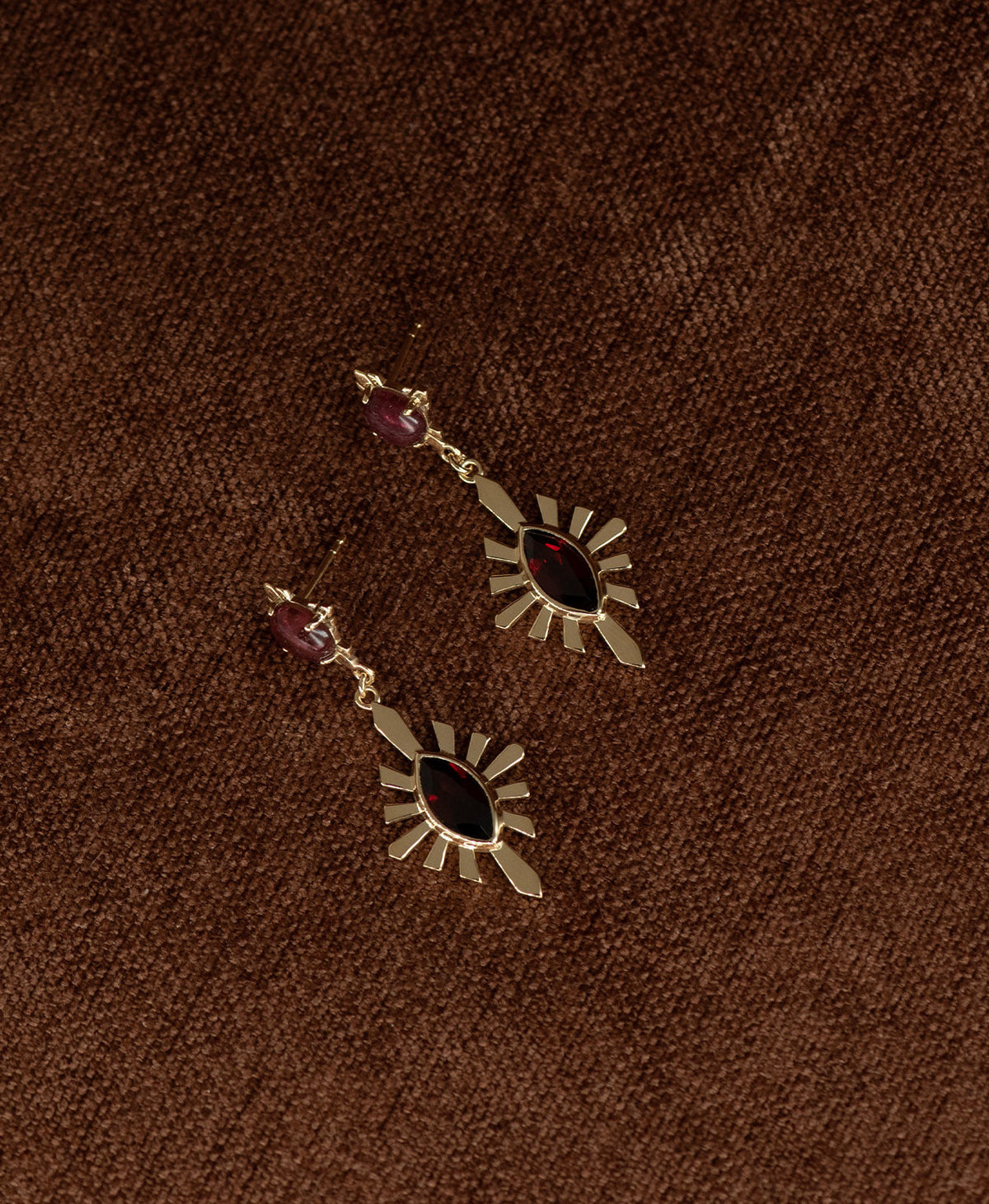 A pair of gold Sisterhood Earrings with a cabochon cut pink Tourmaline, set within a linear border, paired with a marquise cut Garnet nestled with a sunburst frame, displayed against a textured brown fabric background.