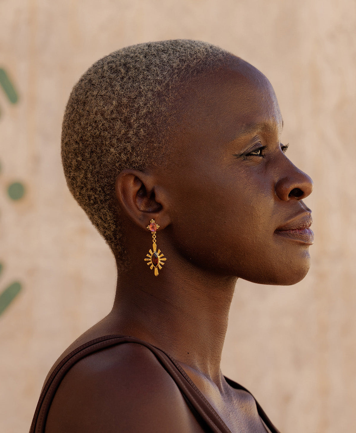 A side profile of a person wearing the  Sisterhood Earring, featuring a cabochon cut pink Tourmaline is set within a linear border, paired with a marquise cut Garnet with a sunburst frame.