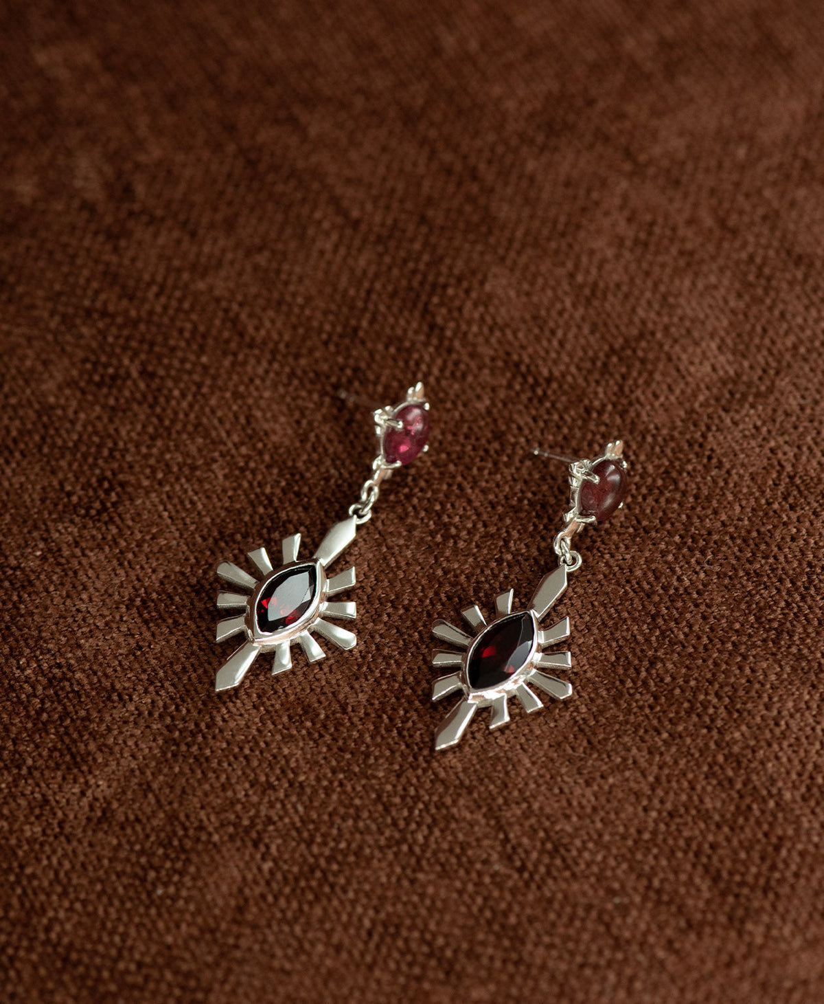 A pair of silver Sisterhood Earrings with a cabochon cut pink Tourmaline, set within a linear border, paired with a marquise cut Garnet nestled with a sunburst frame, displayed against a textured brown fabric background.
