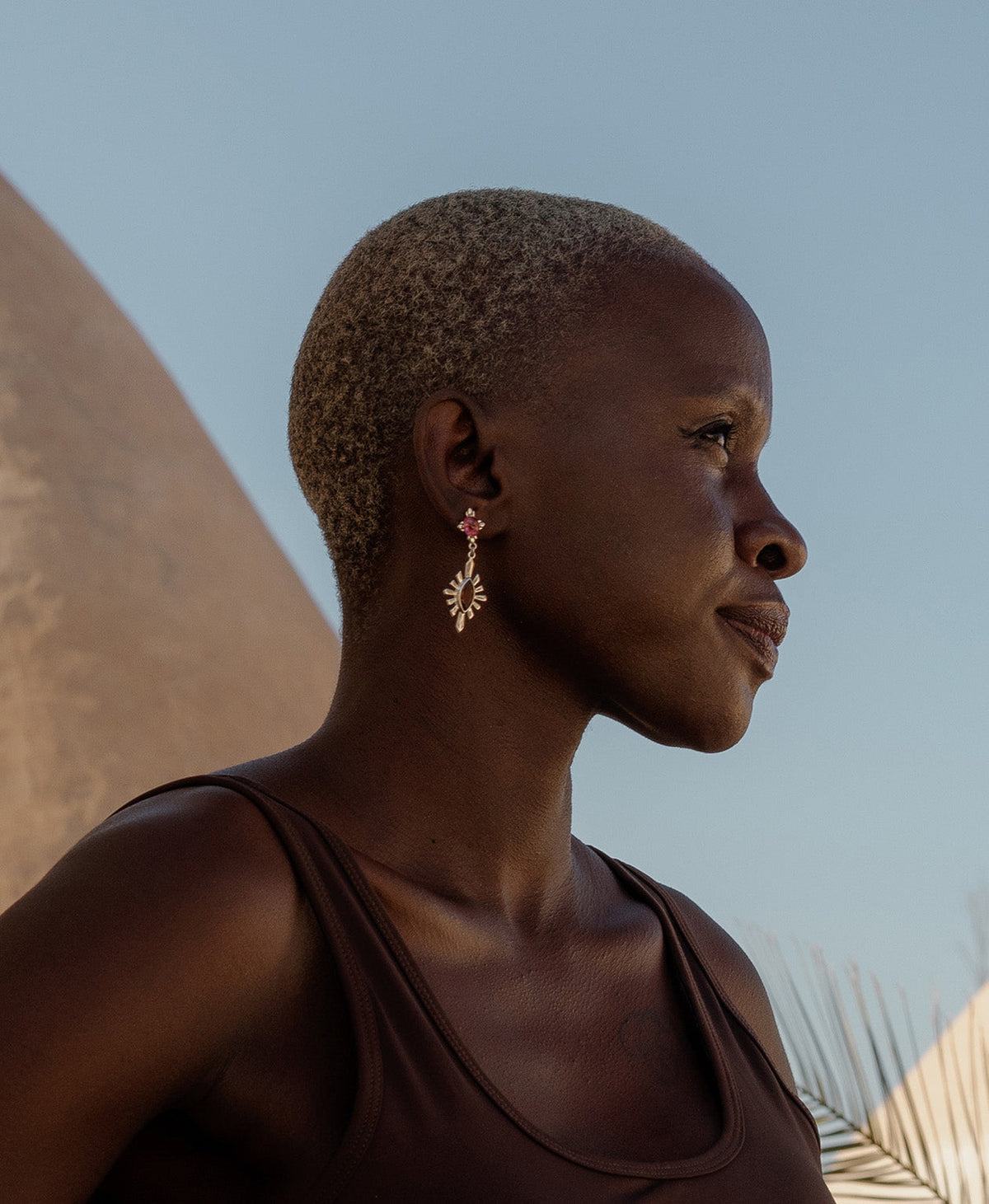 A side profile of a person wearing the silver Sisterhood Earring, featuring a cabochon cut pink Tourmaline is set within a linear border, paired with a marquise cut Garnet with a sunburst frame.