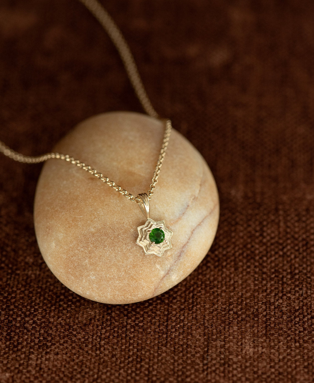 The Sitara Necklace featuring a gold star like pendant with concentric lines that radiate outwards from a Chrome Diopside resting at its centre, displayed over a cream stone on a textured brown fabric background.