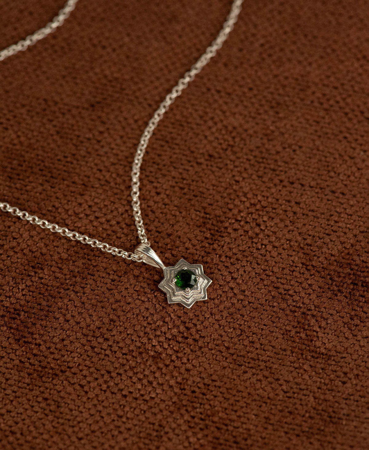Close up of the Sitara Necklace featuring a silver star like pendant with concentric lines radiating outwards from a Chrome Diopside resting at its centre, displayed on a textured brown fabric background.