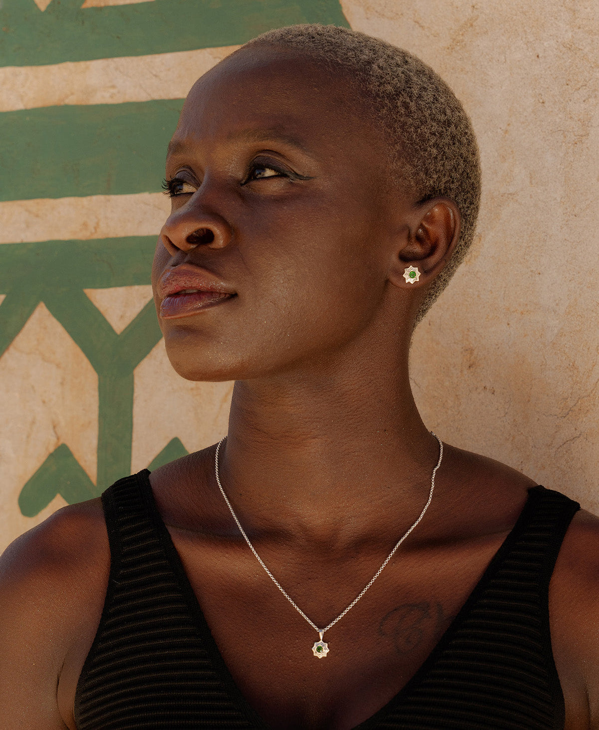 A close-up of a neck wearing the Sitara Necklace, a silver star-like pendant with a central green stone, paired with the matching silver studs. 