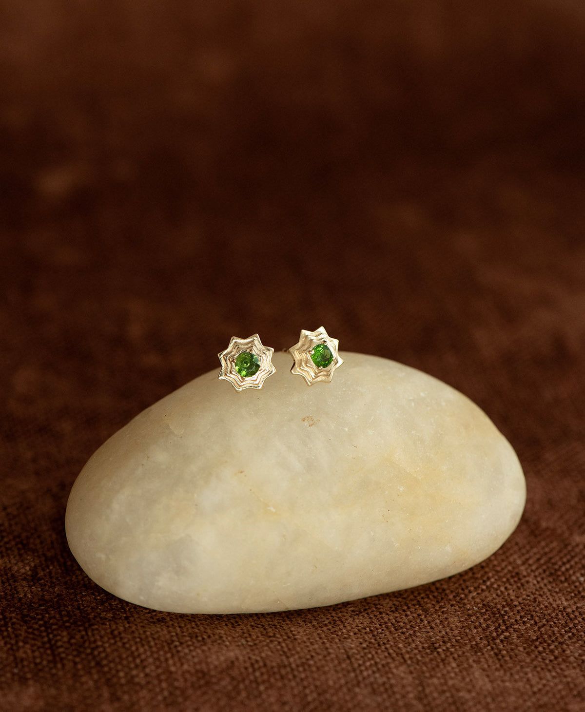 A pair of gold geometric star stud earrings with a central chrome diopside, displayed on a smooth cream coloured stone against a dark brown textured background.