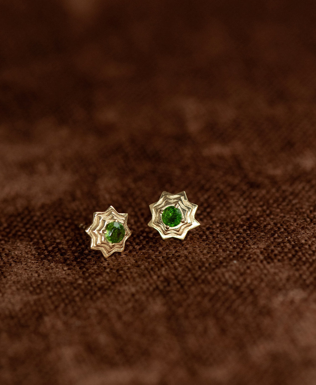 A pair of gold geometric star stud earrings with a central chrome diopside, displayed on a textured brown fabric background.