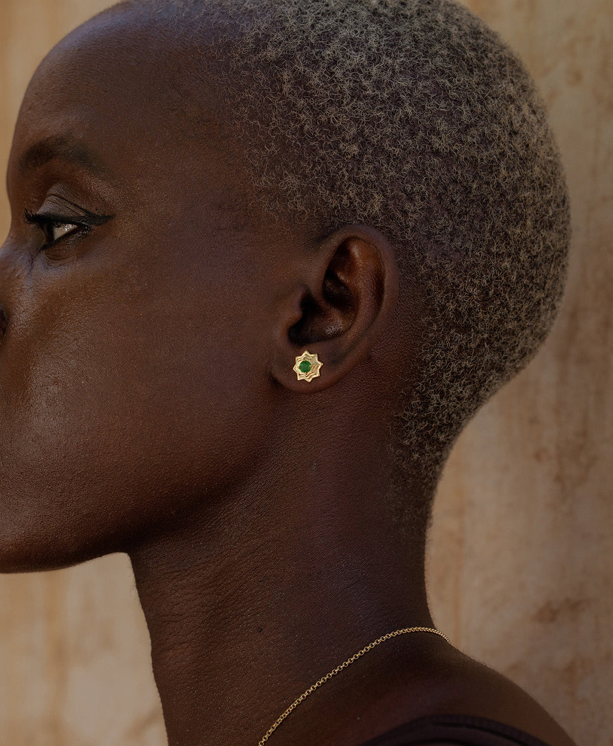 A close-up side profile of a person wearing the Sitara Studs, a gold star-like stud with a central green stone. 