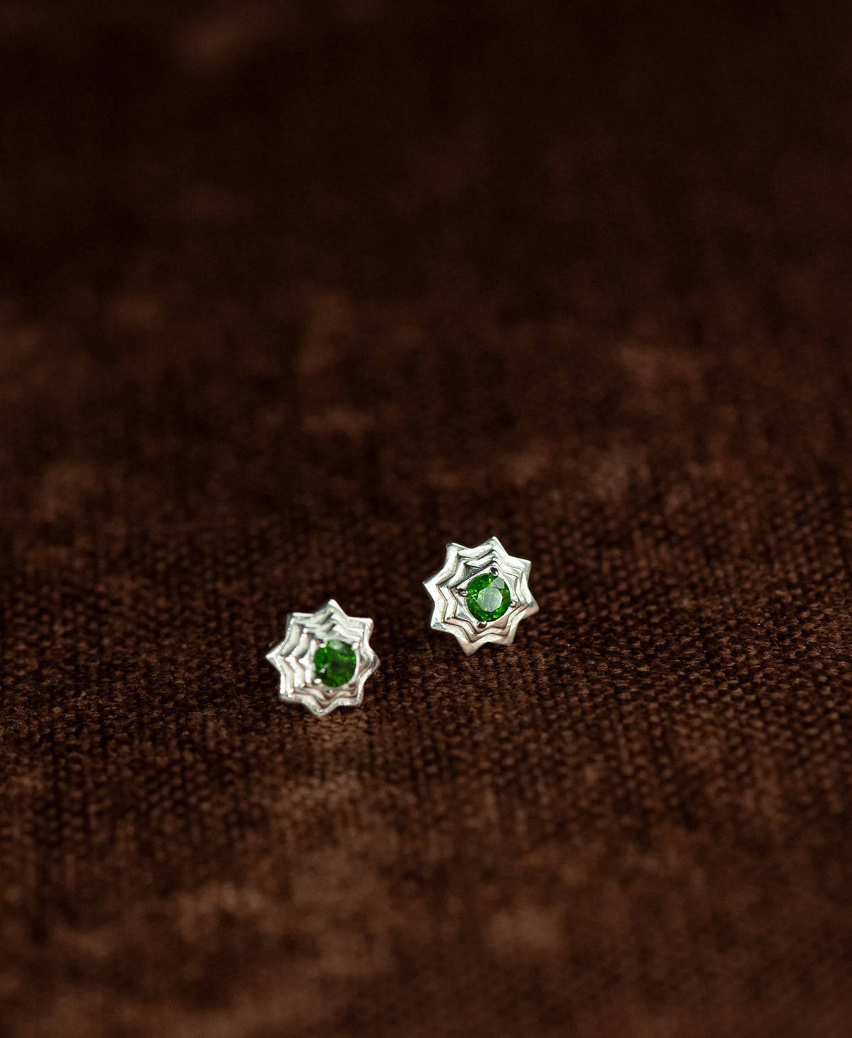 A pair of silver geometric star stud earrings with a central chrome diopside, displayed on a textured brown fabric background.