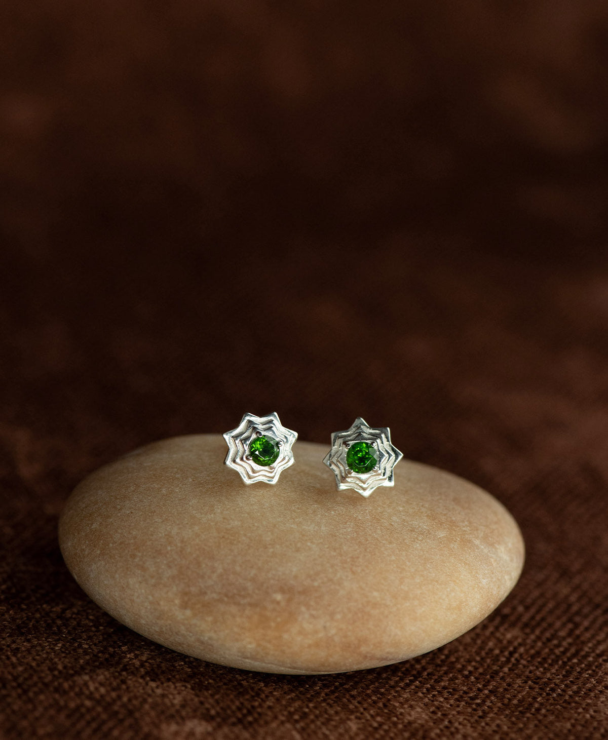 A pair of silver geometric star stud earrings with a central chrome diopside, displayed on a smooth cream coloured stone against a dark brown textured background.