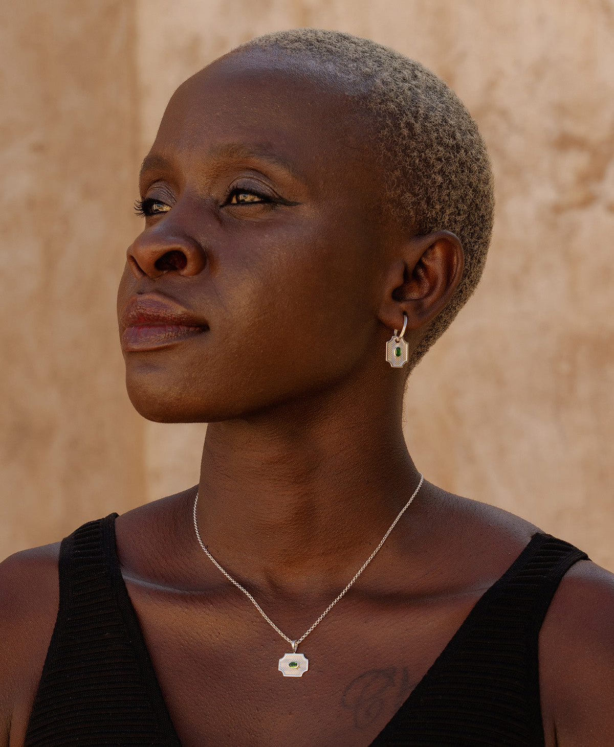 A close-up of a person wearing the Soul Sister Hoops, featuring a textured silver pendant with a central green stone on a hoop., paired with a silver pendant necklace.