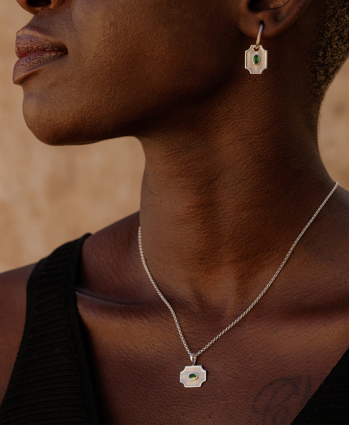 A close-up of a neck wearing the Soul Sister Necklace, a silver textured pendant with a central green stone, suspended from a fine belcher chain, paired with matching silver earrings. 