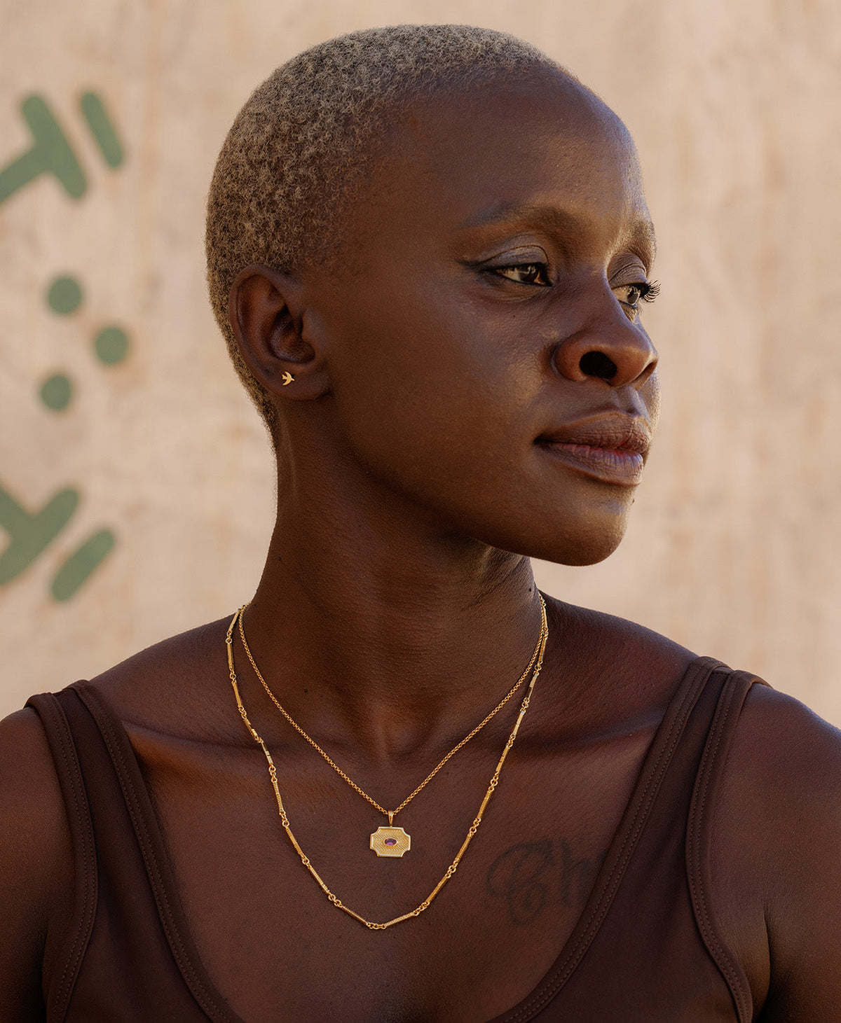 A person wearing the Soul Sister Necklace, a gold textured pendant with a central pink stone, suspended from a fine belcher chain, paired with  longer chain necklace and small gold studs. 